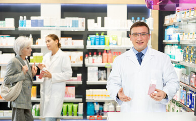 Polite middle-aged male pharmacist demonstrating preparation in box in chemist's shop with large assortment