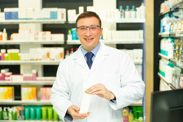 Polite middle-aged male pharmacist demonstrating preparation in box in chemist's shop with large...