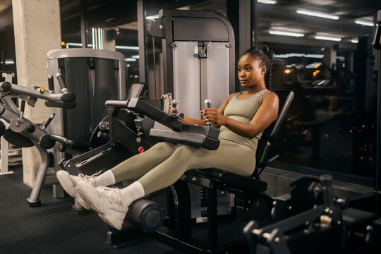 A strong black sportswoman doing workouts at seated leg curl machine at gym.