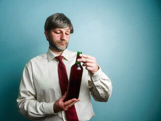 Office worker on blue wall background holding open bottle of red wine. Drinking after work habit for stress relief concept. Model in his 40s with grey beard and hair, wearing light shirt and tie.