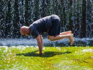 Bald man doing workout with his own body weight in a stream by a waterfall. Male in his 40s with...