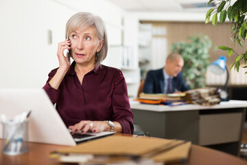 Senior woman clerical worker sitting at table in front of laptop and talking on phone. Her male colleague sitting in background. - obrazy, fototapety, plakaty