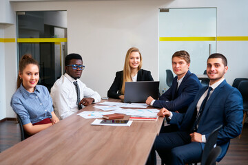 Multiracial corporate team in meeting at office desk, engaged in strategy session with laptop, papers, reflecting teamwork, innovation in modern work environment with focus on diversity and inclusion.