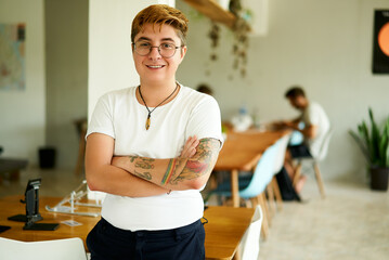 Confident transgender male with tattoos stands in a modern office, smiling colleagues working in the background. He embraces diversity, inclusion in the workplace, wearing casual attire and glasses.