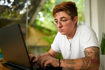 Transgender male concentrates on freelance work from home, using laptop, glasses on, tattoo visible. Gender diversity in remote work setup, inclusive workplace ambiance with home plants.