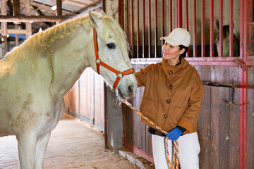 Asian woman horse breeder standing in horse barn and stroking white horse.