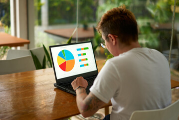 Transgender employee analyzes data for project on laptop at modern workspace. Focused pro prepares...
