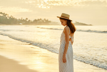 Elegant woman strolls on sunny beach in summer, white polka-dot dress flowing, straw hat on....