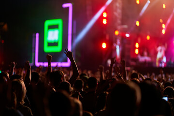 Concertgoers raise their hands, enjoying an energetic live music performance at night. Bright stage lights illuminate the vibrant fest while fans dance and cheer in a packed outdoor venue.