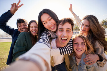 Group of diverse happy young friend taking selfie together looking smiling at camera. Millennial people sharing cheerful moments posing piggyback with peace gestures raised. Friendship and community