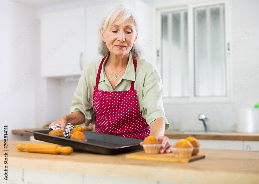 Wall mural Positive old woman in apron taking out just baked cupcakes. Senior woman cooking cakes at home.