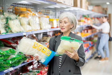 Senior woman in supermarket buys daily groceries, pasta Chinese noodles. Female pensioner buyer...