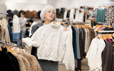 Pleased mature woman choosing short fur coat in clothing shop with large assortment