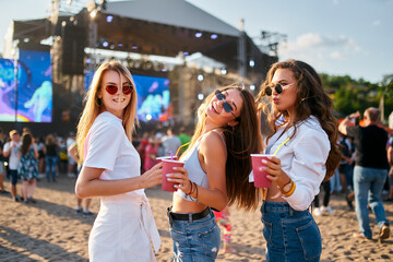 Group of smiling young women with sunglasses enjoy summer music festival on beach holding colorful...