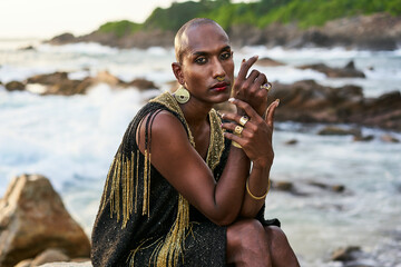 Queer black person in luxury dress, jewelry sits on rocks in ocean. Lgbtq ethnic fashion model...