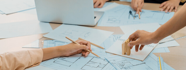 Professional architect hand draws a blueprint on table with architectural document and wooden block scatter around at office. Design and Planing concept. Focus on hand. Closeup. Delineation.