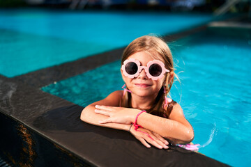 Smiling girl in pink sunglasses rests on pool edge in summer. Child enjoys sunny day at water park....