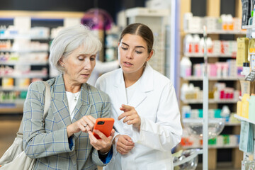 Female pharmacist and elderly woman customer reading prescription together on smartphone screen in...