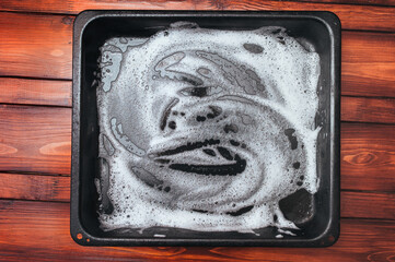 White foam and soap on a black baking sheet that stands on a wooden table. Abstract pattern. The concept of washing the greasy surface of a frying pan.