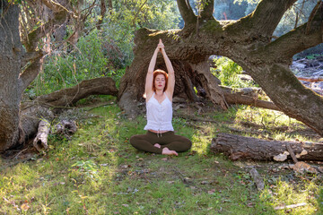 young redhead sexy woman doing asana yoga exercises lotus pose with hands and arms up in nature in sportswear in the sun