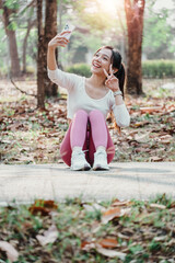 Cheerful young woman sitting on a path in a sunlit park takes a selfie, flashing a peace sign.