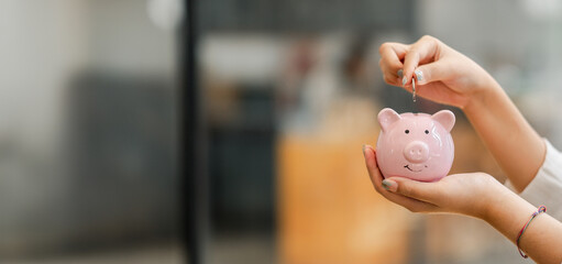 Hands inserting a coin into a pink piggy bank, symbolizing the concept of savings and financial...