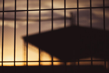 Silhouette of an industrial building behind a lattice fence against sunset yellow orange sky in the...