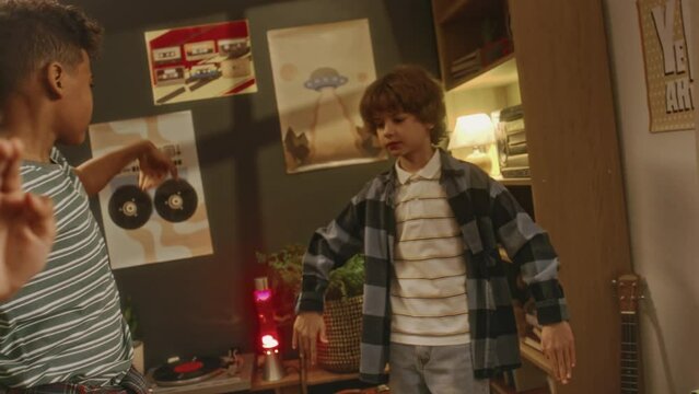 Pan handheld footage of two diverse best friends playing music on beatbox player and performing robot dance and waving dance while hanging out after school in retro styled house, nostalgic 90s