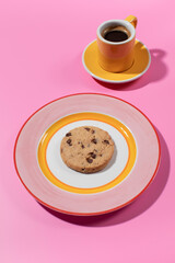 Chocolate chip cookie on a bright plate, pink backdrop with a cup of coffee.