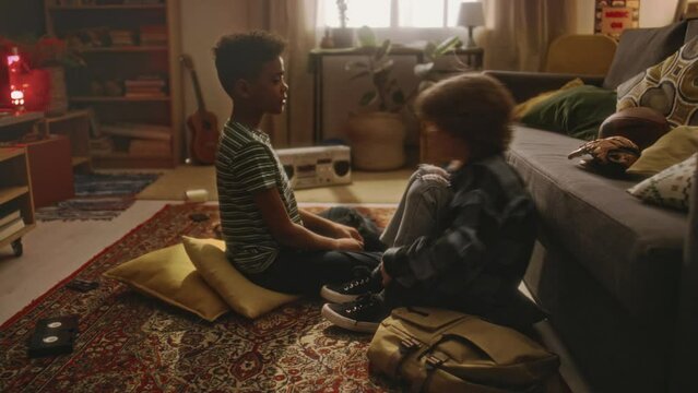 Side full footage of little Caucasian boy sharing secret with his African American best friend, boy giving oath to keep secret by raising left hand and drawing cross on heart in retro living room 90s