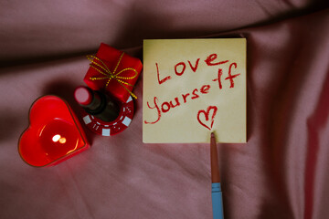 A note to self with a call to love yourself drawing with red lipstick on a silk dark pink cloth flatly. A burning heart-shaped red candle, gift box. Happy Valentines Day 2024. Love yourself first. - obrazy, fototapety, plakaty
