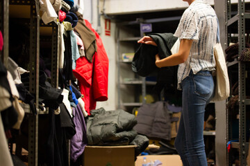 Young woman among rows of shelves of shabby clothes. A girl picks out clothes at a refugee shelter,...