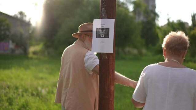 An elderly couple tapes a missing dog notice to a pole while standing outdoors in a city park on a sunny summer day. An upset married couple leaves, holding hands