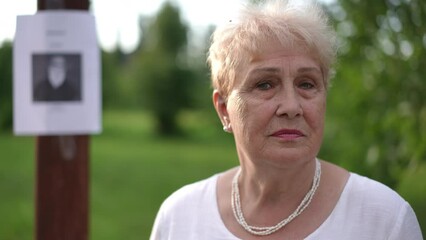 In the foreground, an elderly upset woman looks sadly at the camera while standing in a city park outdoors on a summer day. In the background, out of focus, is a notice on a pole about a missing