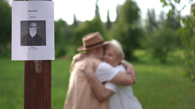 A happy elderly couple hugs, smiles and kisses while standing on the grass in a city park on a summer day against the backdrop of a missing elderly man notice post