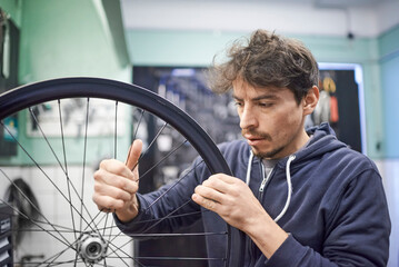 Young hispanic man checking the tension of the spokes of a bicycle wheel in his bike workshop. Real...