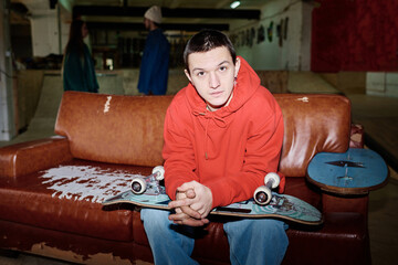 Confident gen Z guy sitting on leather couch with skateboard on lap in skatepark looking at camera,...