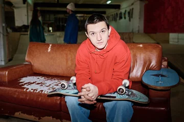 Fotobehang Skateboard Confident gen Z guy sitting on leather couch with skateboard on lap in skatepark looking at camera, copy space  © AnnaStills