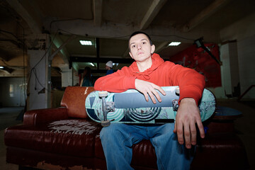 Naklejka premium Flash photo of self-confident gen Z man wearing red hoodie and jeans sitting on shabby couch in skatepark looking at camera