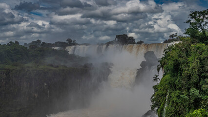 A picturesque tropical waterfall. White foaming streams collapse from the rocks into the river. Spray and fog rise into the air. Lush green vegetation. Clouds in the blue sky. Iguazu Falls. Argentina