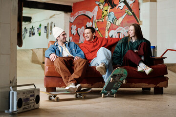 Group of three modern male and female friends hanging out in skatepark sitting on couch, chatting...