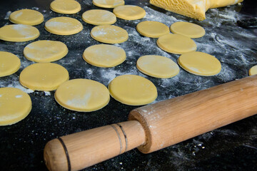 photo of a preparation of simple cookies with many already cut out in the shape of a circle, out of focus wooden roll and large dough, on a black table with a lot of flour
