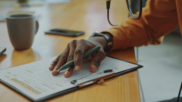 Hands of male podcast host sitting at desk in recording studio, proofreading content plan and putting marks on paper. Close-up view