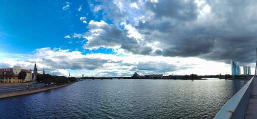 Panoramic cityscape over water, with dramatic clouds and a bridge in the distance.