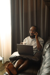African American man freelancer talking on mobile phone and using laptop in living room