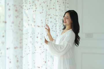 Asian woman in white pajamas is opening the curtains and looking out the window smiling in her room in the morning. Soft sunlight. Vacation, lifestyle concept.