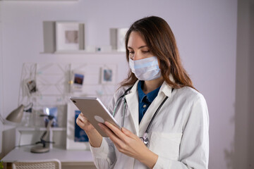 A female doctor in a mask and white uniform uses a tablet in her work. Technologies
