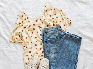 Polka dot muslin blouse, blue jeans, sneakers - beautiful comfortable urban style women's clothing on a light background, top view