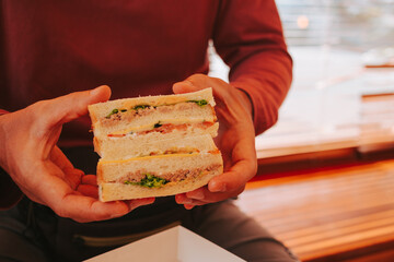 A man holds a double sandwich with tuna and vegetables with his hands on the background of a window. Delicious toast for breakfast in the morning