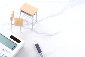 学校の机と椅子、電卓がイメージする、高騰を続けて家計を圧迫する教育費のイメージ
